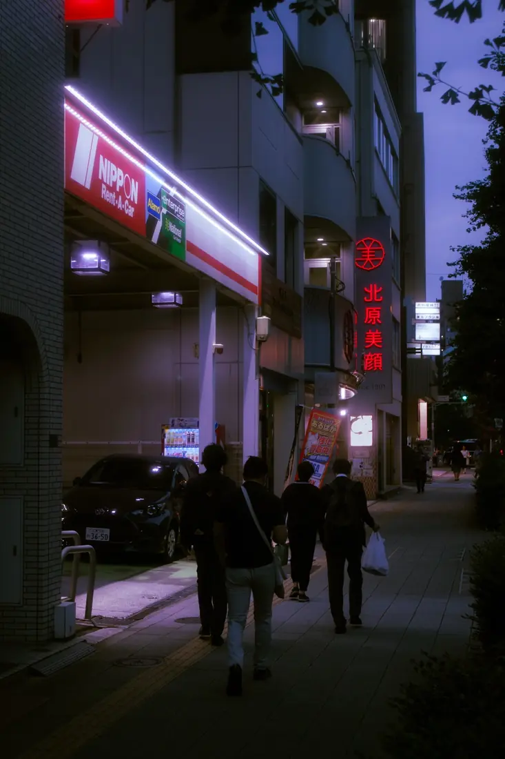 Pedestrians walking beneath red neon and lavender evening light