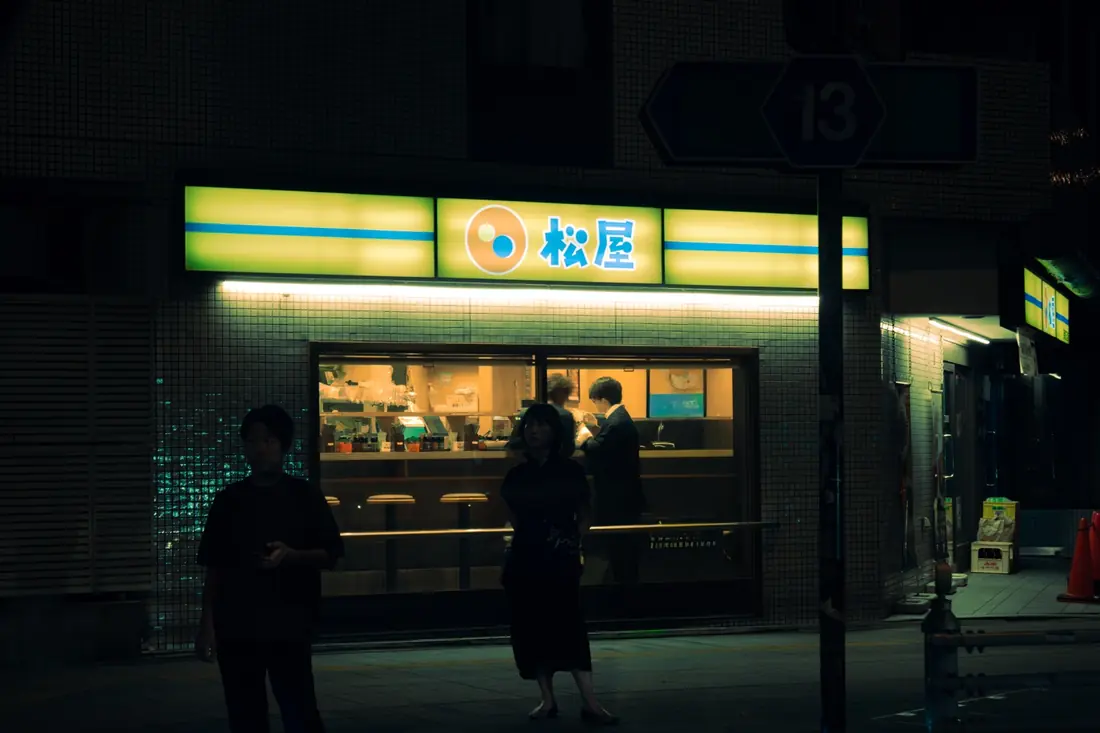Figures gathered outside a warmly lit late-night restaurant